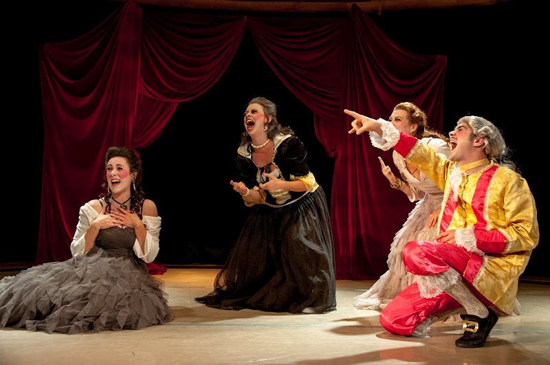 Quatre personnages en costumes du XVIIe siècle éclatent de rire devant un rideau rouge